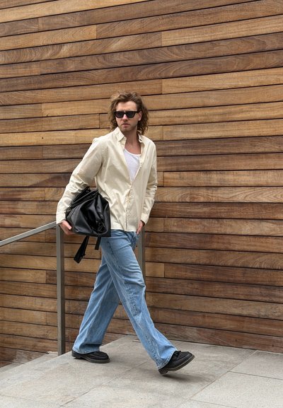 Hombre con gafas de sol, camisa beige, camiseta blanca y jeans azules camina cerca de una pared de madera, llevando un bolso negro sobre el hombro.