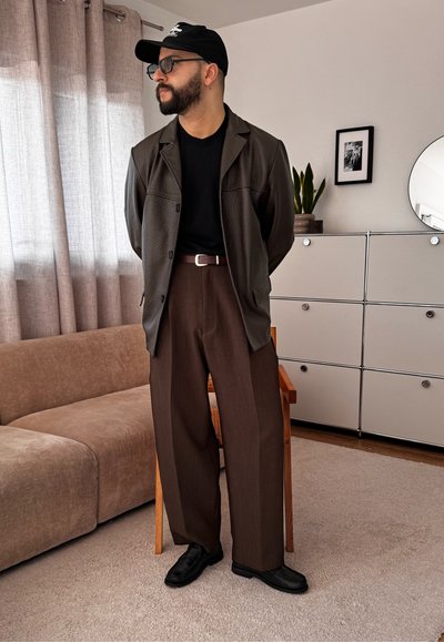 Hombre barbudo con gorra negra, gafas, chaqueta de cuero marrón oscuro, camisa negra, pantalones anchos marrones y zapatos negros en el interior.