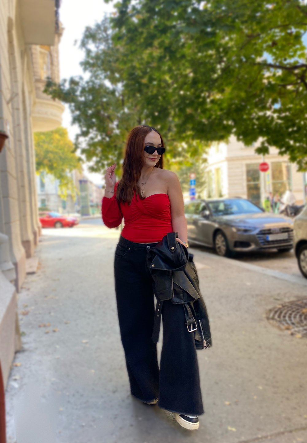 Κόκκινη τοπ με έναν ώμο, μαύρα φαρδιά τζιν και μαύρη δερμάτινη ζακέτα. Τα αξεσουάρ περιλαμβάνουν γυαλιά ηλίου και χρυσές αλυσίδες. Εξωτερικό περιβάλλον.