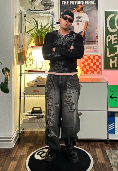 Camisa negra de manga larga con diseño intrincado; jeans grises destrozados y holgados; botas de plataforma negras; gafas de sol; detalle de cadena de plata.