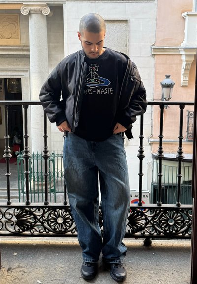 Chaqueta bomber negra, camiseta gráfica negra con "ANTI-WASTE", jeans azul desgastados y zapatillas negras. De pie en un balcón con barandillas.