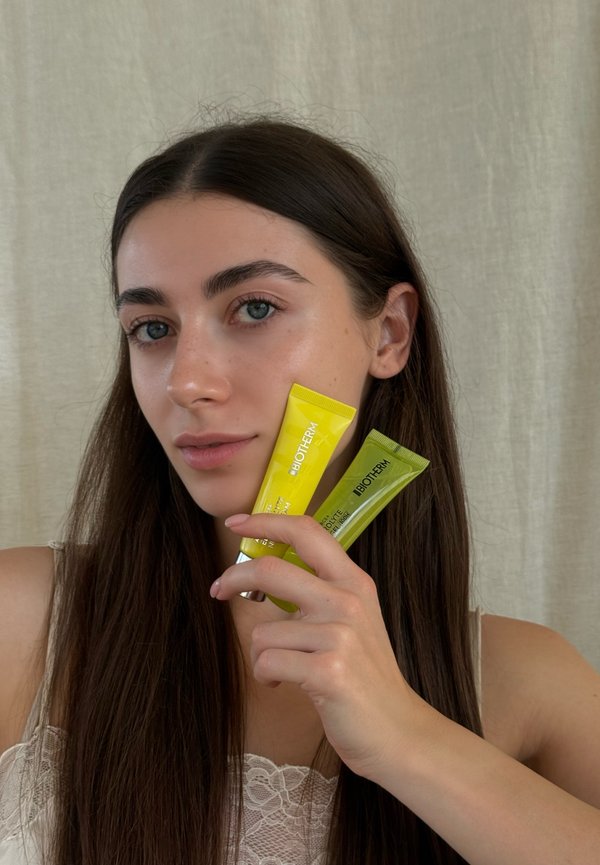 Una mujer sostiene dos tubos de productos para el cuidado de la piel de Biotherm junto a su cara, uno amarillo y otro verde, delante de una pared lisa.