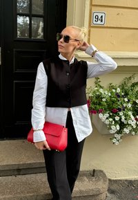 Μαύρο κοντό γιλέκο πάνω από άσπρο πουκάμισο, συνδυασμένο με μαύρα παντελόνια. Κρατάει μια κόκκι�νη τσάντα. Γυαλιά ηλίου και λουλουδάτο φόντο.