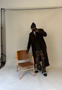 Muž v tmavě hnědém kabátu, červené kravatě, maskáčových kalhotách, černé čepici a slunečních brýlích stojí u dřevěné židle na jednoduchém studiovém pozadí.