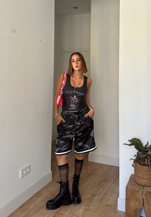 Top negro sin mangas con diseño gráfico, pantalones cortos de camuflaje con ribetes blancos, botas de combate negras y un bolso rosa. Incluye collares en capas y calcetas transparentes.