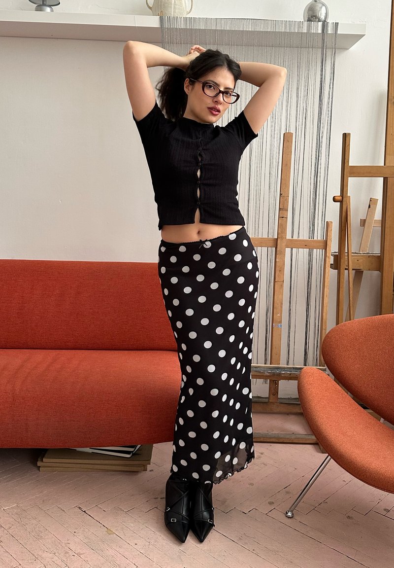 Une femme portant des lunettes, un haut noir court et une jupe noire à pois se tient entre un canapé orange et un fauteuil dans un studio.