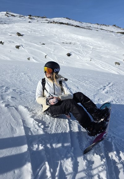 Persona con casco negro y gafas de protección sentada sobre la nieve con una tabla de snowboard, con una montaña nevada y un cielo azul despejado de fondo.