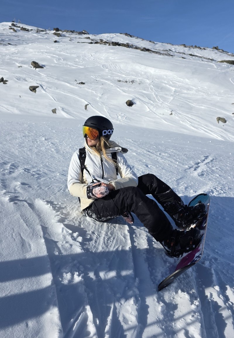Personne portant un casque noir et des lunettes de protection assise sur la neige avec un snowboard, une montagne enneigée et un ciel bleu clair en arrière-plan.