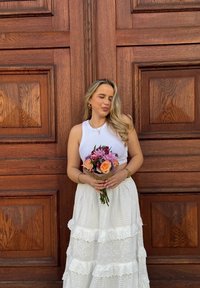 Weißes Crop-Top, kombiniert mit einem geschichteten, weißen Rock mit Spitzenbesatz. Sie hält einen Strauß aus pinken und lila Blumen vor einem Holz-Hintergrund.