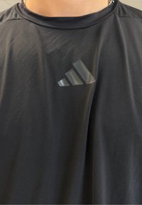 Melna sporta krekls ar spīdīgu Adidas logo kreisajā krūtī, kas uzvilkts cilvēkam ar sudraba ķēdes kulonu.