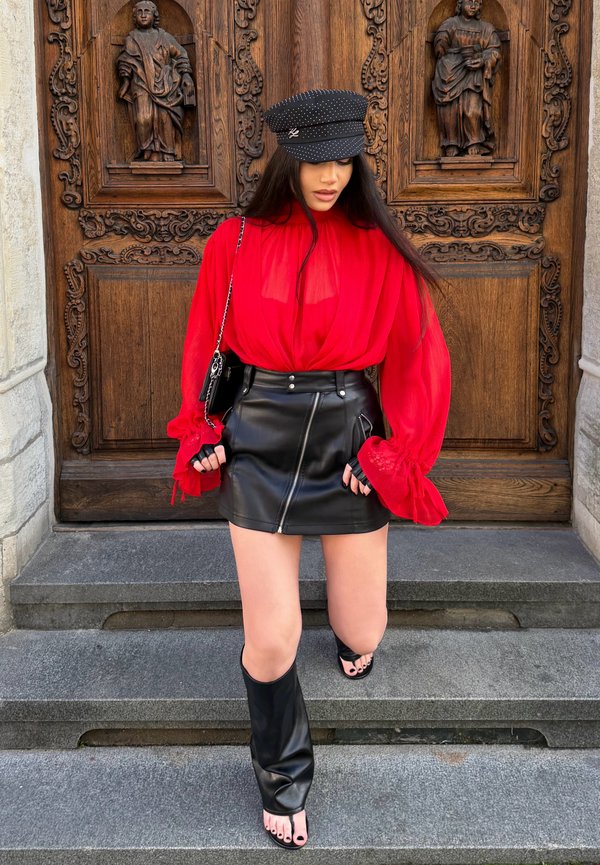 Une femme vêtue d’un chemisier rouge, d’une jupe en cuir noire, d’une casquette et de bottes pose sur des marches en pierre devant une porte en bois richement sculptée.