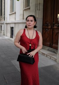 Donna in un vestito rosso senza maniche che tiene una borsa nera e degli occhiali da sole, in piedi su un marciapiede cittadino vicino a una porta di legno.