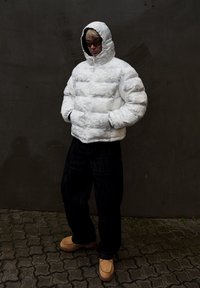 Hvit vattert jakke med geometrisk mønster, hette og glidelås, kombinert med mørke vide bukser og beige slip-on sko.