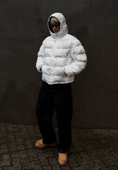 Chaqueta blanca acolchada con patrón geométrico, con capucha y cremallera, combinada con pantalones anchos oscuros y zapatos beige sin cordones.