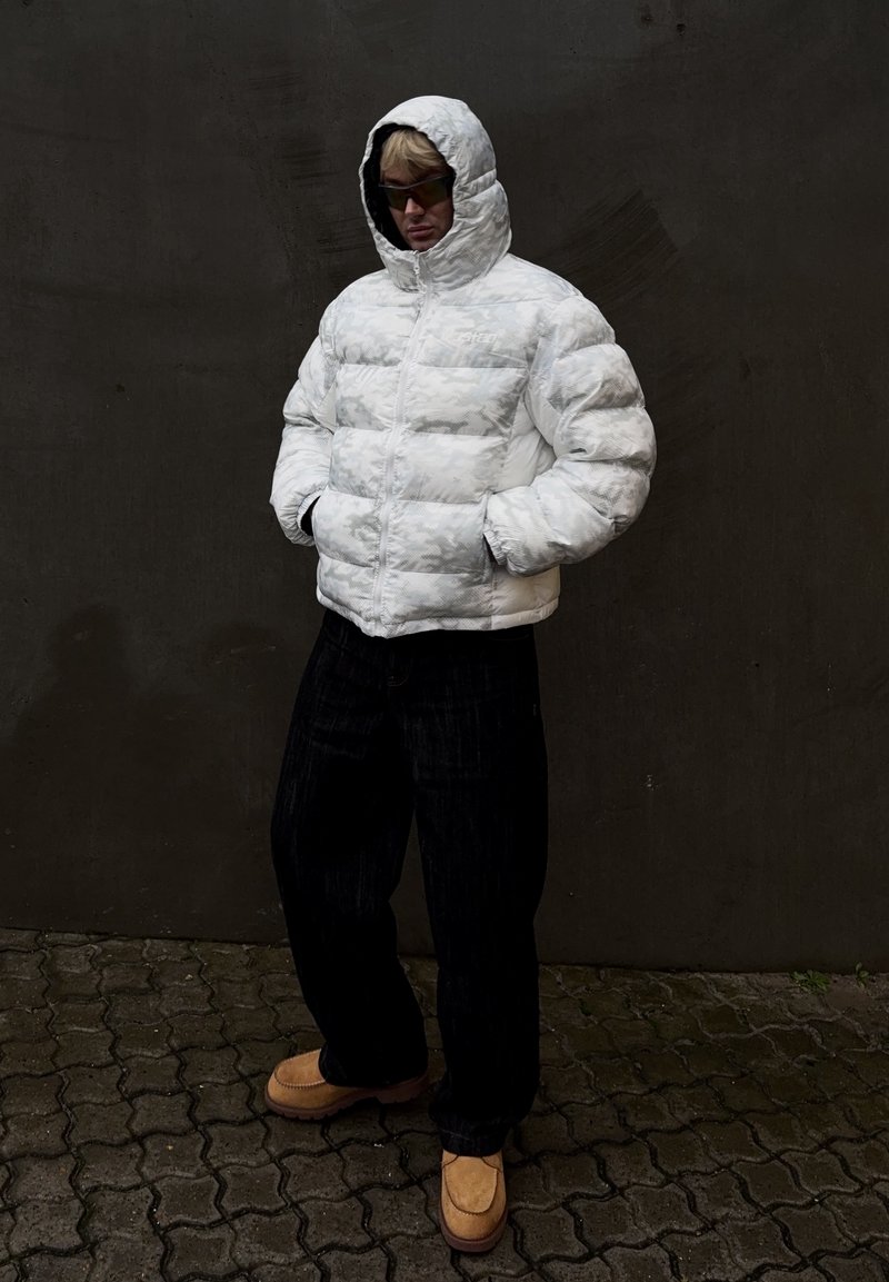 Giacca bianca imbottita con motivo geometrico, con cappuccio e cerniera, abbinata a pantaloni larghi scuri e scarpe beige slip-on.