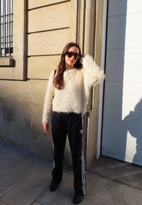 Pälsig vit tröja, svarta träningsbyxor med vita ränder, svarta skor, solglasögon, stående mot en stenmur. Naturligt ljus.