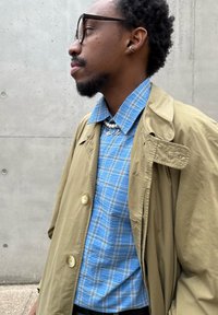 Άνδρας που φοράει μπεζ παλτό τύπου trench, μπλε πουκάμισο με καρό σχέδιο, κολιέ με μαργαριτάρια και γούρια, στρογγυλά γυαλιά και ασημένιο σκουλαρίκι στο αυτί, στέκεται μπροστά σε έναν τσιμεντένιο τοίχο.