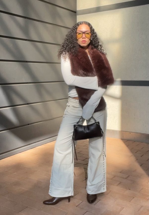 Mulher com cabelo cacheado, usando óculos de sol amarelos, colete de pelagem castanho, blusa branca de manga longa, calças flare brancas, segurando uma bolsa preta, em pé à luz do sol.