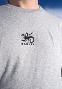 Gros plan sur une chemise grise avec un logo de scorpion noir et le texte « OAKLEY » brodé sur la poitrine, sur fond de ciel bleu.