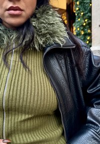 Geacă neagră de piele cu guler verde de blană, peste un top verde măsliniu cu model striat. Prezintă un fermoar argintiu care coboară pe centru.