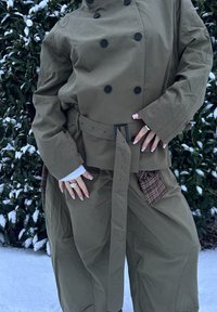 Veste oversize vert olive avec boutons croisés, taille ceinturée et pantalon à jambes larges avec détail de poche à carreaux. Fond enneigé.