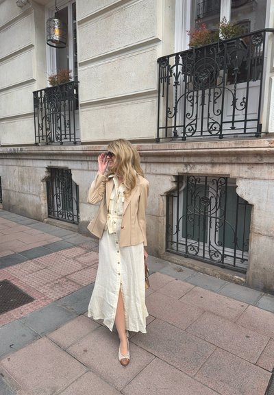 Chaqueta beige ajustada sobre un vestido blanco con botones y una abertura hasta el muslo, combinado con zapatos planos beige y un bolso pequeño.