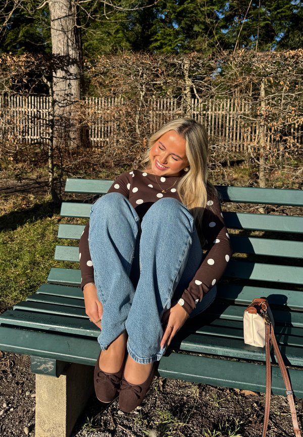 Joven con cabello rubio, ropa de lunares y jeans sentada con las piernas cruzadas, sonriendo en un banco verde de parque con una bolsa a su lado.