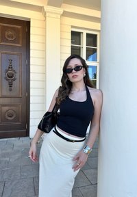 Black halter neck top, cream skirt with black belt, black handbag; silver jewellery; sleek sunglasses; standing by a white column and wooden door.