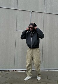 Felpa nera con zip e tasca, abbinata a pantaloni cargo beige larghi e sneaker bianche. Gli accessori includono occhiali da sole e un foulard nero.