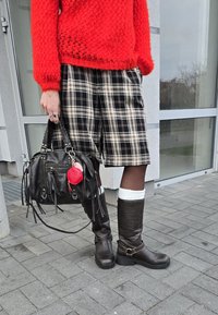 Schwarze Lederhandtasche mit Fransen, roter Schlüsselanhänger, roter Strickpullover, schwarz-beige karierte Shorts, hohe braune Lederstiefel und weiße Socken.