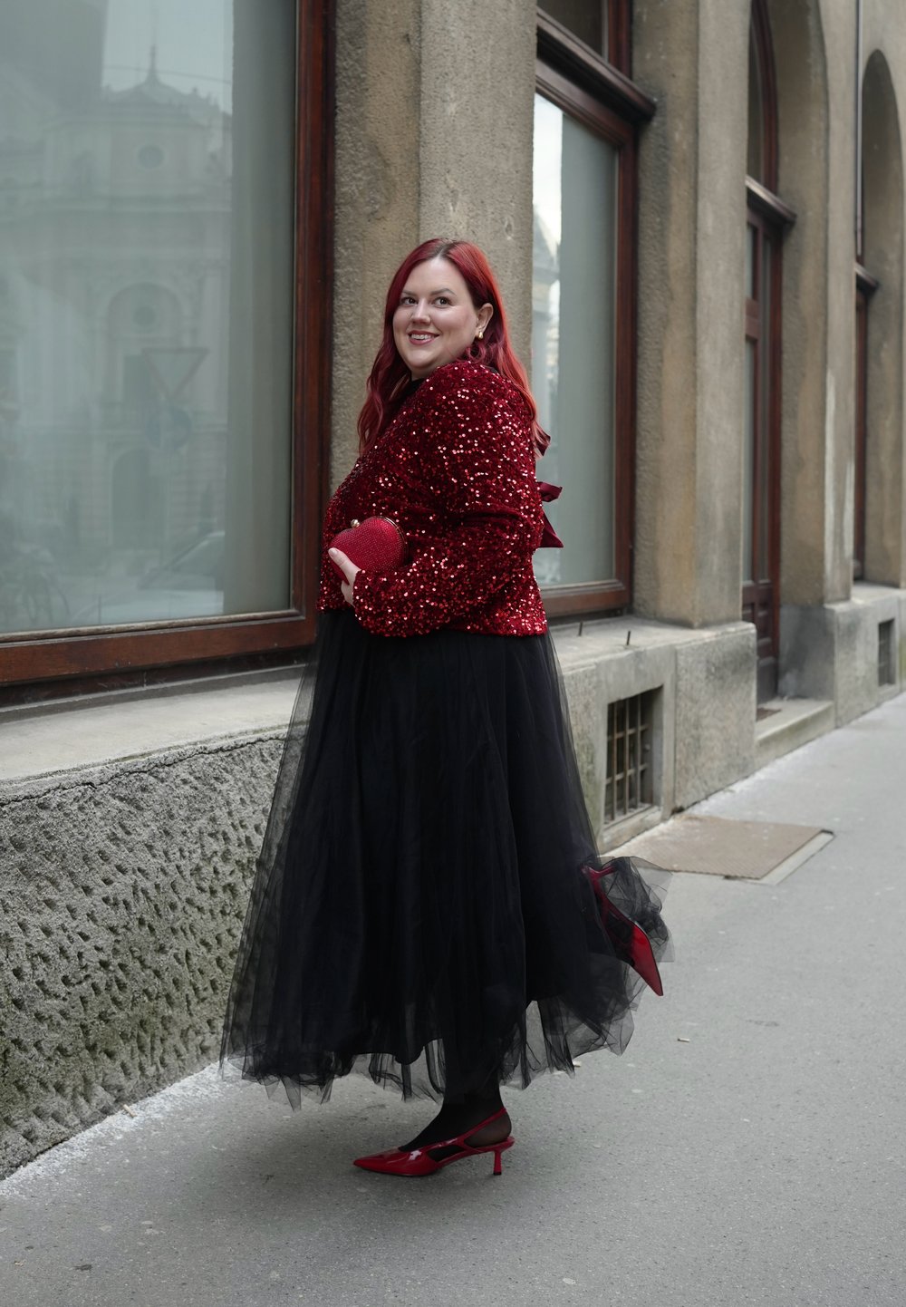 Une femme aux cheveux roux porte une veste rouge à sequins, une jupe en tulle noire et des talons rouges, tenant une pochette rouge assortie sur un trottoir en ville.