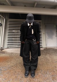 Homme portant un manteau noir, un pantalon large noir, des bottes noires, une casquette noire, des colliers superposés, debout sur du gravier sous le auvent d'un bâtiment.