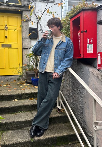 Joven con chaqueta de mezclilla y suéter bebiendo café, apoyado en una barandilla junto a unas escaleras de piedra cerca de una puerta amarilla y una caja de extintor de incendios roja.