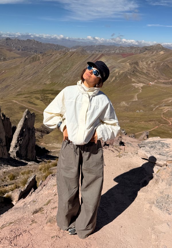 Personne portant des lunettes de soleil, une casquette noire, une veste blanche et un pantalon gris, debout avec les mains sur les hanches sur un sentier de montagne rocheux sous un ciel clair.