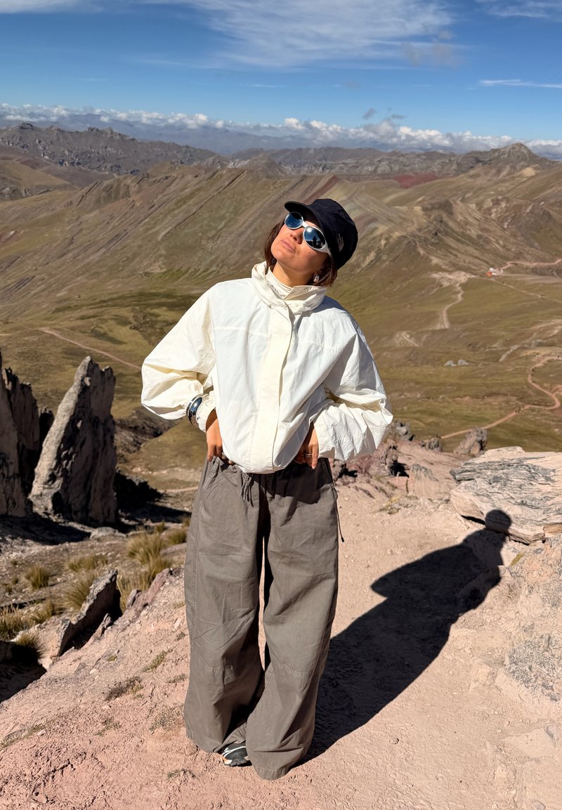 Personne portant des lunettes de soleil, une casquette noire, une veste blanche et un pantalon gris, debout avec les mains sur les hanches sur un sentier de montagne rocheux sous un ciel clair.