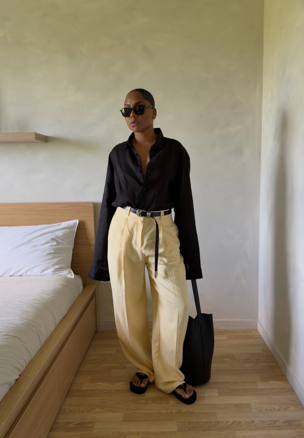 Persona con camisa negra, pantalones anchos beige, cinturón negro, gafas de sol negras y sandalias negras, de pie en un dormitorio minimalista sosteniendo una bolsa tote negra.