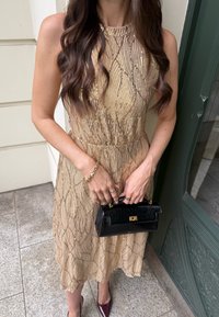 Mujer con un vestido dorado de lentejuelas que sostiene un pequeño bolso negro, lleva una pulsera dorada, anillos y zapatos puntiagudos de color rojo oscuro.