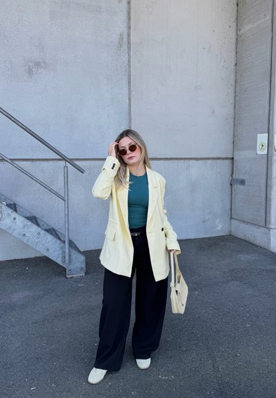 Mujer con gafas de sol, blazer amarillo pálido, top azul verdoso, pantalones anchos negros, zapatos blancos y que sostiene un bolso beige, está de pie junto a unas escaleras de cemento.