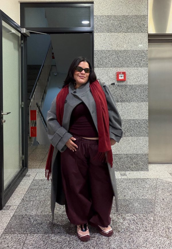 Une femme portant des lunettes de soleil, un manteau gris, une écharpe rouge et un pantalon ample bordeaux pose dans un couloir carrelé, à côté d’une porte ouverte donnant sur la cage d’escalier.
