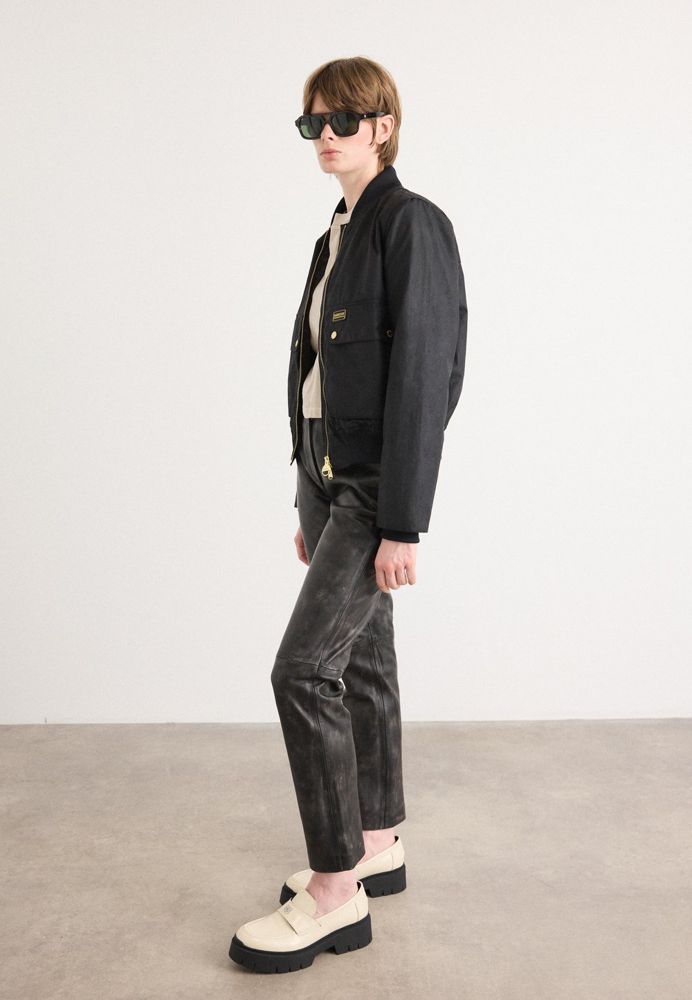 Casaco bomber preto com ferragens douradas, blusa bege, calças de pele preta estampadas e sapatos com sola grossa em creme. O modelo usa óculos de sol escuros.