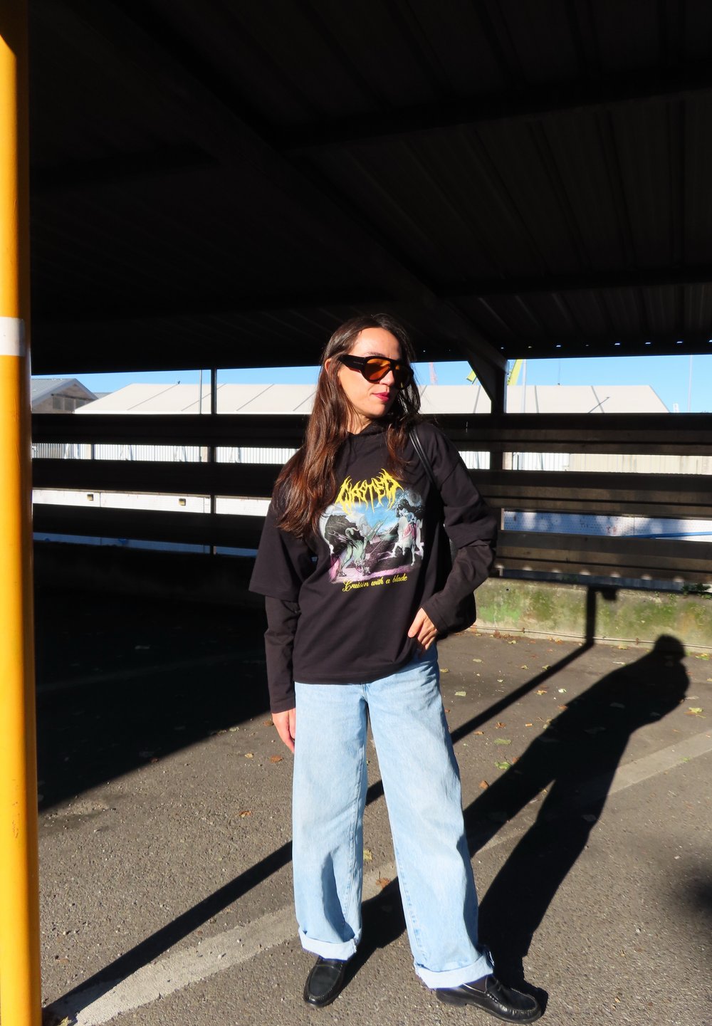 Čierna grafická mikina s farebným dizajnom, svetlomodré široké džínsy a čierne topánky. Slnečné okuliare a dlhé vlasy dotvárajú outfit.