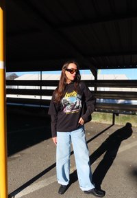 Čierna grafická mikina s farebným dizajnom, svetlomodré široké džínsy a čierne topánky. Slnečné okuliare a dlhé vlasy dotvárajú outfit.