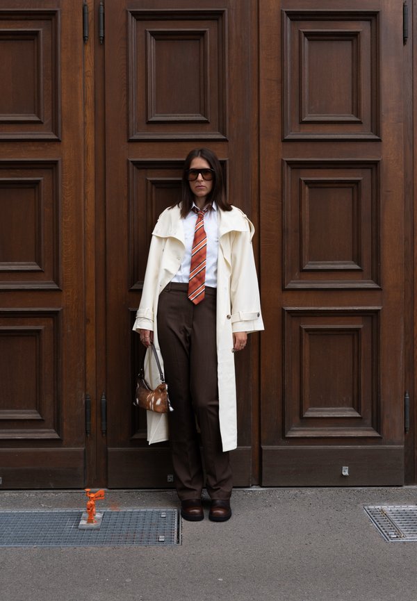 Mujer con pantalones marrones, camisa blanca, corbata naranja a rayas y abrigo largo color crema, de pie junto a grandes puertas de madera con paneles, sosteniendo un pequeño bolso marrón.