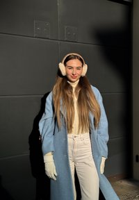 Jeune femme aux longs cheveux portant un manteau bleu clair, des gants blancs, un col roulé crème, un pantalon blanc et un cache-oreilles beige, debout contre un mur sombre.