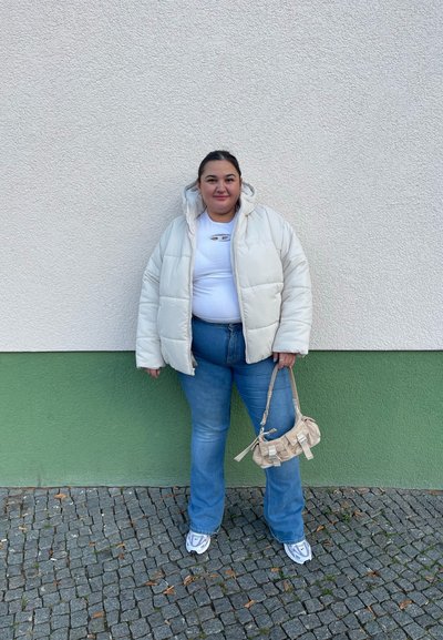 Chaqueta blanca acolchada, top blanco entallado, jeans azules de pata de elefante, bolso beige de hombro con asas y zapatillas blancas con detalles grises. Fondo de adoquines grises.