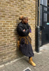 Sort bomberjakke, svart skjørt med brune frynser, brune støvler. Tilbehør inkluderer store øredobber og briller, med håret stylet i en knute.