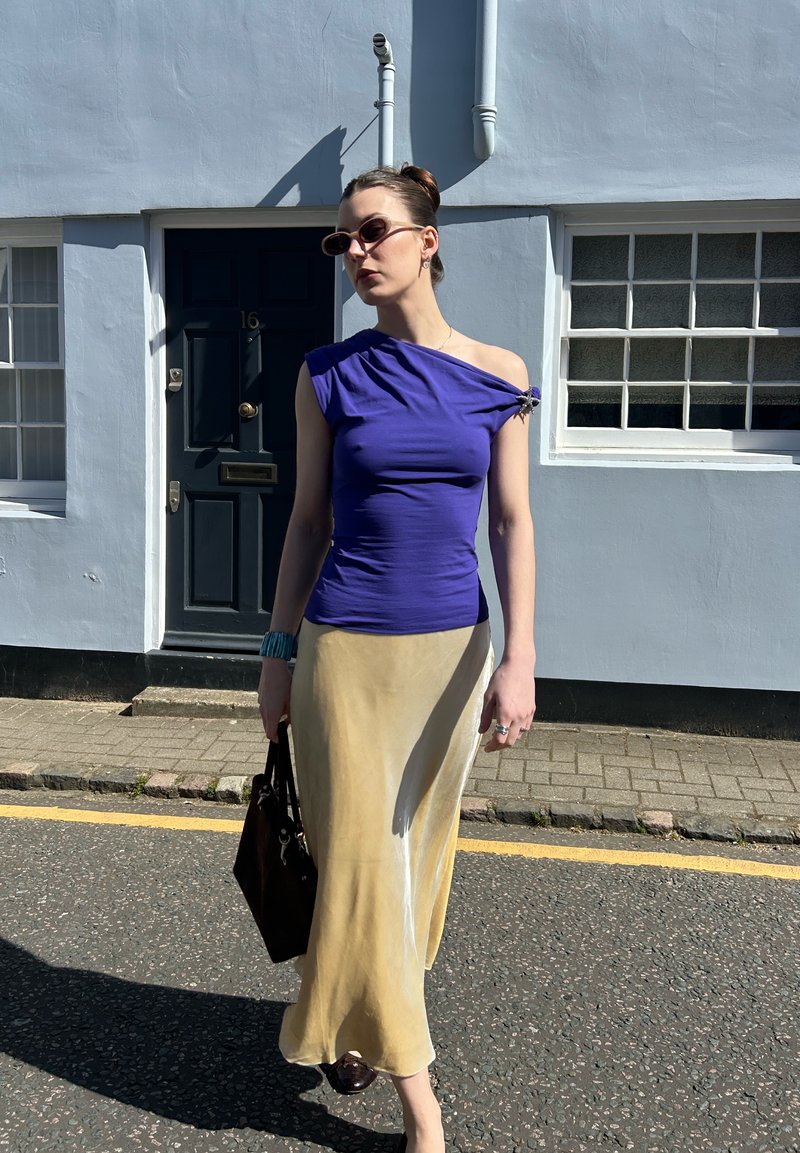 Donna con top viola a una spalla e gonna beige, tiene una borsa nera, cammina su una strada con un edificio bianco e una porta scura sullo sfondo.