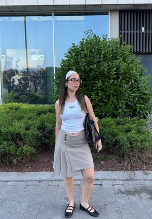 Top sin mangas blanco con logo, falda plisada gris con botones, zapatos planos negros y una diadema de lunares. Fondo: follaje verde.