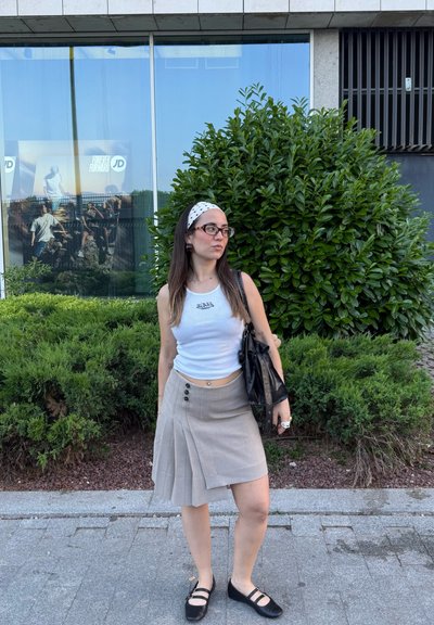 Top sin mangas blanco con logo, falda plisada gris con botones, zapatos planos negros y una diadema de lunares. Fondo: follaje verde.