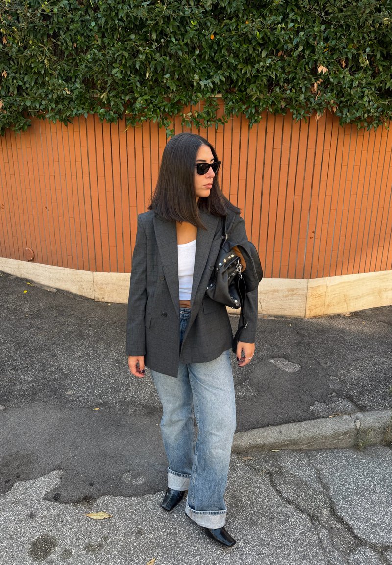 Blazer cinzento aos quadrados sobre uma camisola branca, combinado com jeans azuis claros de perna larga e botas pretas de salto. Mala preta com tachas prateadas.